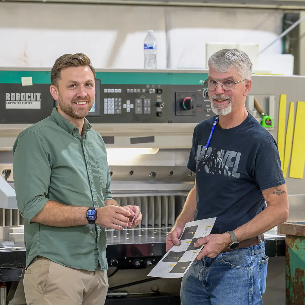 Two Koke Printing team members reviewing a printed sheet beside a paper cutter.