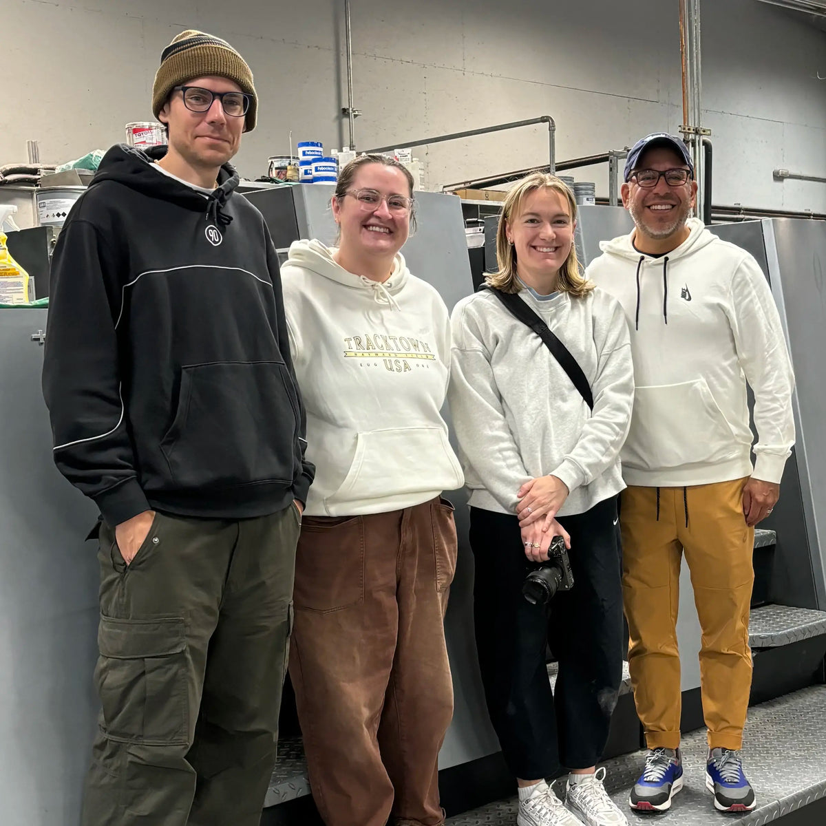 Four visitors posing beside a printing press at Koke Printing, one wearing a TrackTown USA sweatshirt.