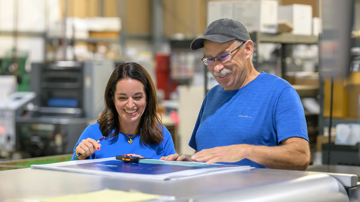Two Koke Printing team members smiling as they check a printed sheet beside a paper cutter.