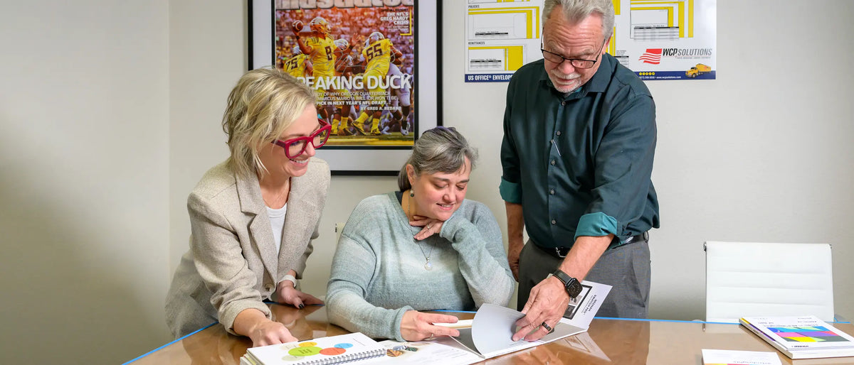 Three people review printed samples and a booklet together at a conference table.