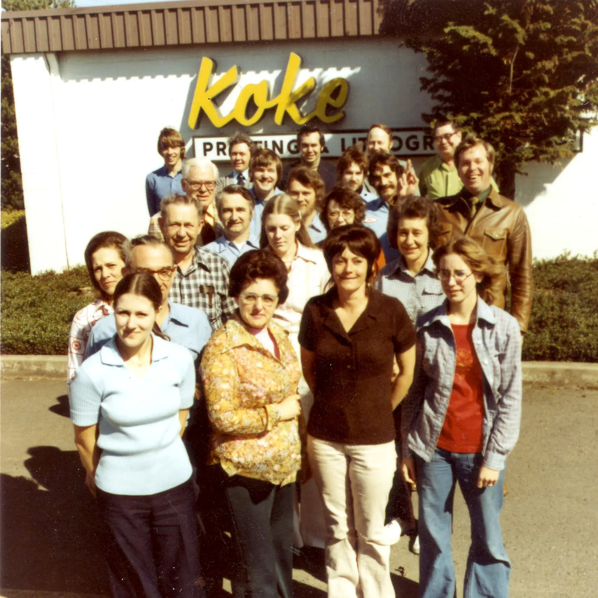 Historic group photo of the Koke staff outside a building beneath the yellow Koke sign.