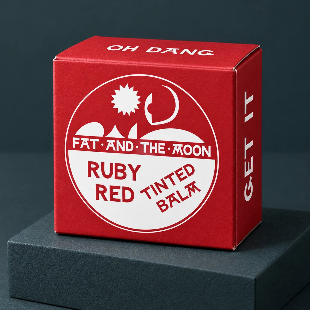 Fat and the Moon Ruby Red Tinted Balm carton showing its front, top, and side on a deep-charcoal studio riser.