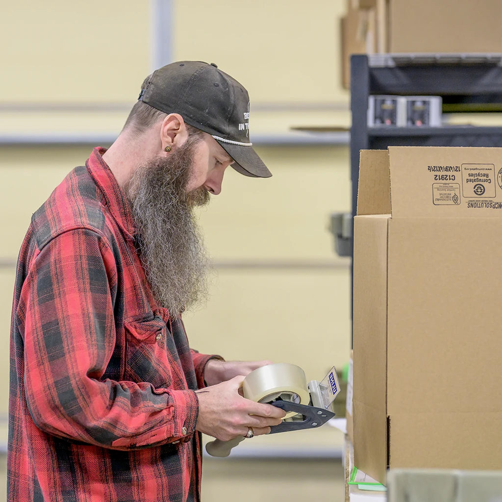 A worker loading a packing tape dispenser beside cardboard shipping boxes.