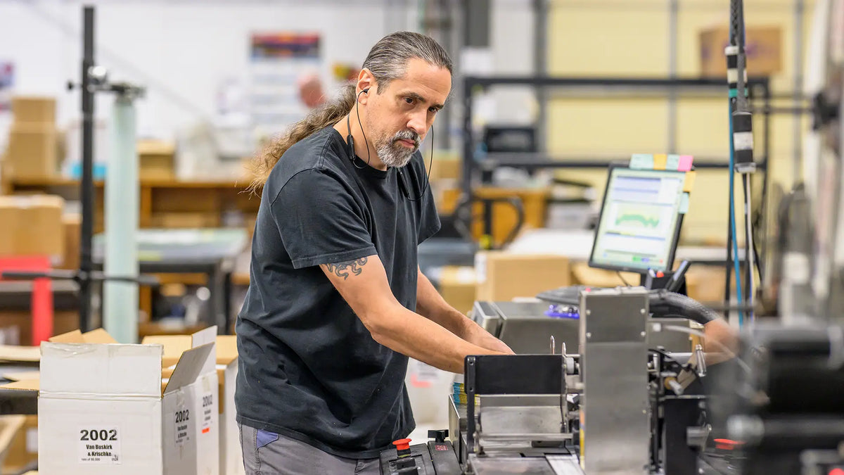 A print shop worker feeding printed pieces into mail processing equipment.