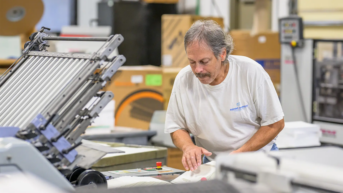 A print shop worker aligning printed pieces at a paper folding machine.