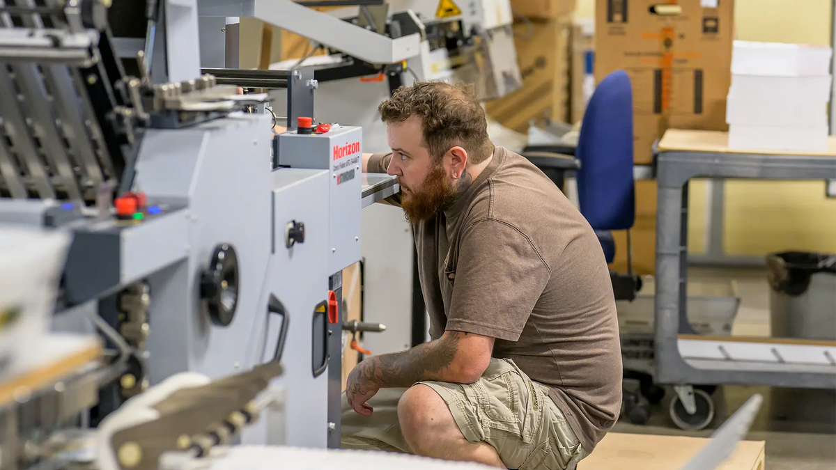 A print shop worker crouching beside a Horizon paper folding machine to check its setup.