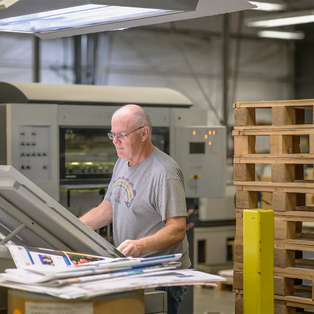 A print shop worker checking printed sheets at a printing press control station.