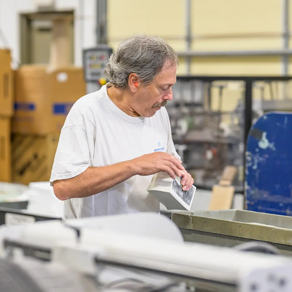 A print shop worker inspecting a stack of printed pieces beside finishing equipment.
